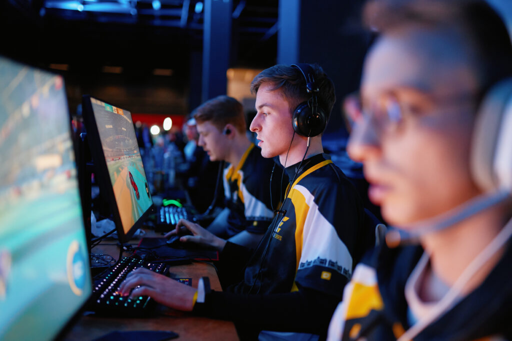Drei Gamer in Teamtrikots spielen mit Headsets an nebeneinanderstehenden PCs bei Rival Rush Finale an der gamescom lan. Illustration zum Thema Erfolgsfaktoren im Gaming Marketing.