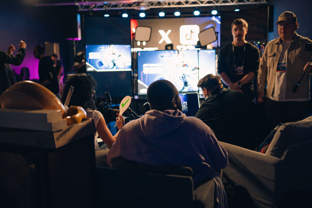 Mehrere Personen sitzen bei einem Gaming Event vor Bildschirmen und verfolgen eine Spielszene. Im Hintergrund sind Monitore, Streaming Lichter und ein grosses Display mit Social Media Symbolen zu sehen. Die Atmosphäre wirkt wie eine Live Gaming Aktivierung mit Publikum, Technik Setup und Event Beleuchtung.