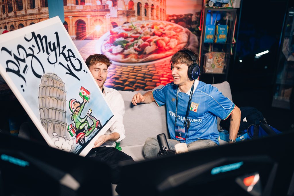 Zwei Personen sitzen im Italien Länderstudio beim YR2V-Finale 2025 auf einem Sofa. Eine hält ein Graffiti Plakat mit dem Schiefen Turm von Pisa, die andere trägt Kopfhörer und ein blaues Trikot.