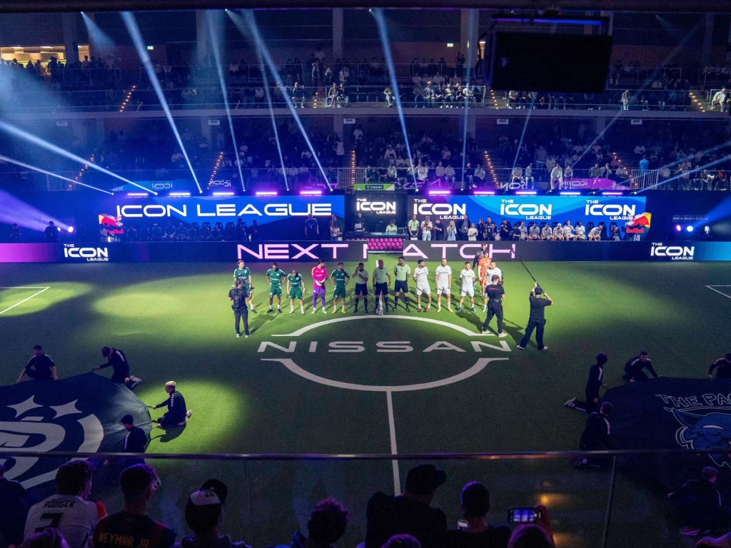 Teams der Icon League stehen vor Spielbeginn auf dem Hallenfussballfeld. Zuschauer*innen auf den Tribünen, Lichtstrahlen und grosse LED-Banden umgeben das Spielfeld.