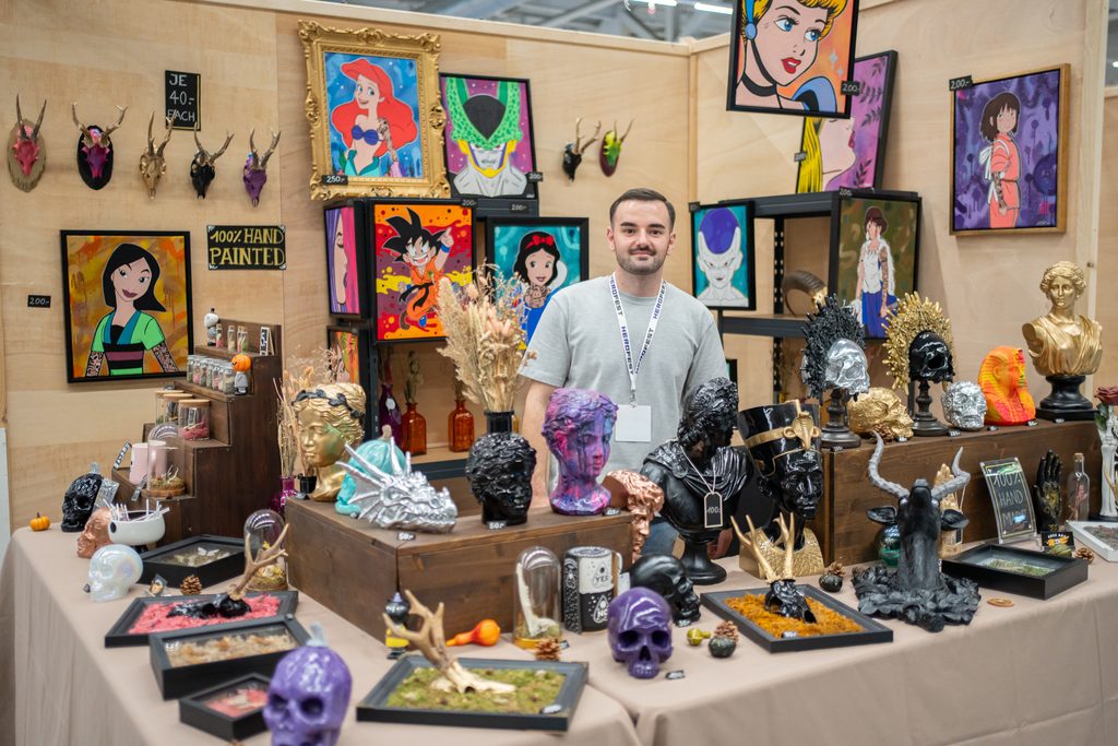 Artist-Stand am HeroFest in Bern mit bunten Gemälden von Anime- und Disney-Figuren sowie dekorativen Büsten und Totenköpfen. Ein Aussteller steht hinter den präsentierten Kunstwerken.