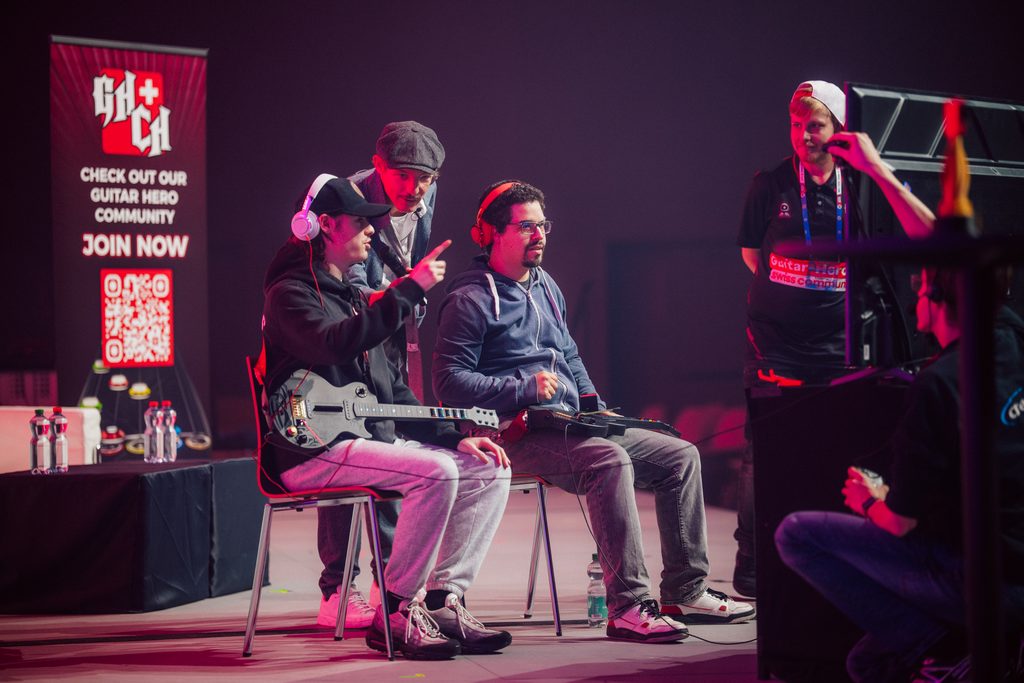 Zwei Personen mit Kopfhörern und Gitarren-Controllern spielen Guitar Hero, weitere schauen zu. Community-Erlebnis als Beispiel für Erfolgsfaktoren im Gaming Marketing.