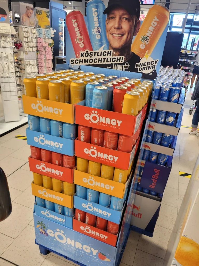 Verkaufsdisplay mit Gönrgy-Energydrinks in blauen, roten und gelben Dosen. Darüber wirbt ein Aufsteller mit einem Porträt von MontanaBlack für die Marke.