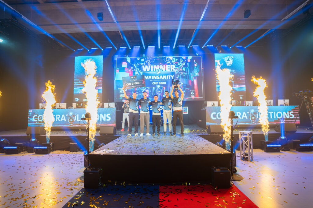 Fünf Mitglieder des Esports-Teams MYINSANITY stehen mit einem Pokal auf einer Bühne. Hinter ihnen verkündet eine große Leinwand ihren Sieg und ein Preisgeld von 3.000 Schweizer Franken. Flammensäulen und Konfetti begleiten die Feier.